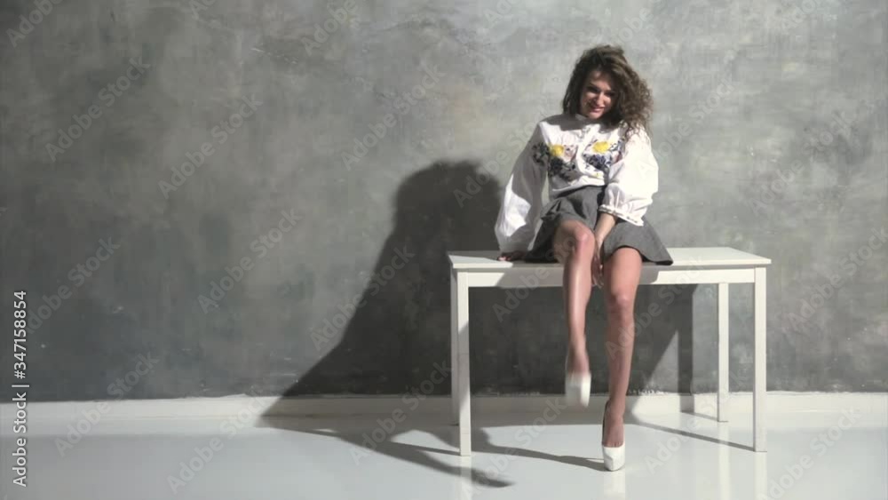 The young beautiful woman with curly hair poses, wriggles and flirts in front of a camera on table, white high heels, a short skirt, a grey background, she coquets, different emotions The young beautiful woman with curly hair poses, wriggles and flirts in front of a camera on table, white high heels, a short skirt, a grey background, she coquets, different emotions