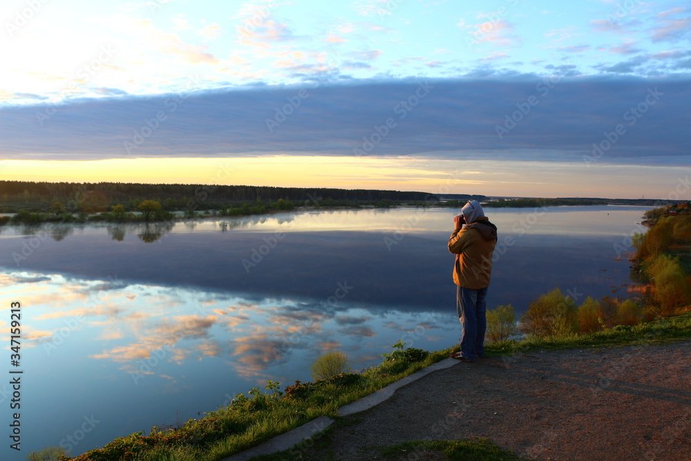 A man in a jacket and hood illuminated at dawn with a camera on a high Bank takes a picture of Golden clouds shining in the river on a blue surface.Reflection of the sky in the water expanses.Russia