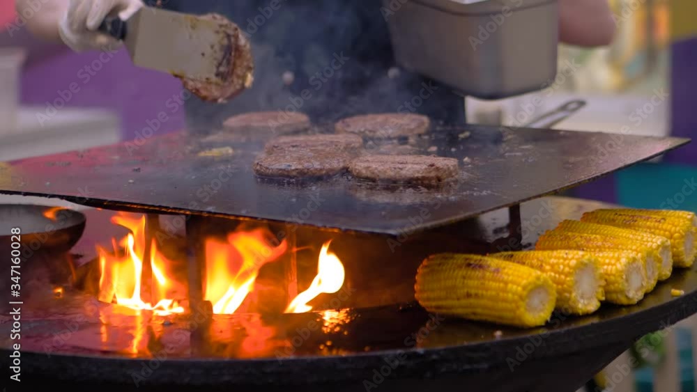 Super slow motion: chef grilling fresh yellow corn cobs and meat cutlets for burgers on brazier with hot flame at summer food market - close up view. Outdoor cooking, gastronomy, street food concept Super slow motion: chef grilling fresh yellow corn cobs and meat cutlets for burgers on brazier with hot flame at summer food market - close up view. Outdoor cooking, gastronomy, street food concept