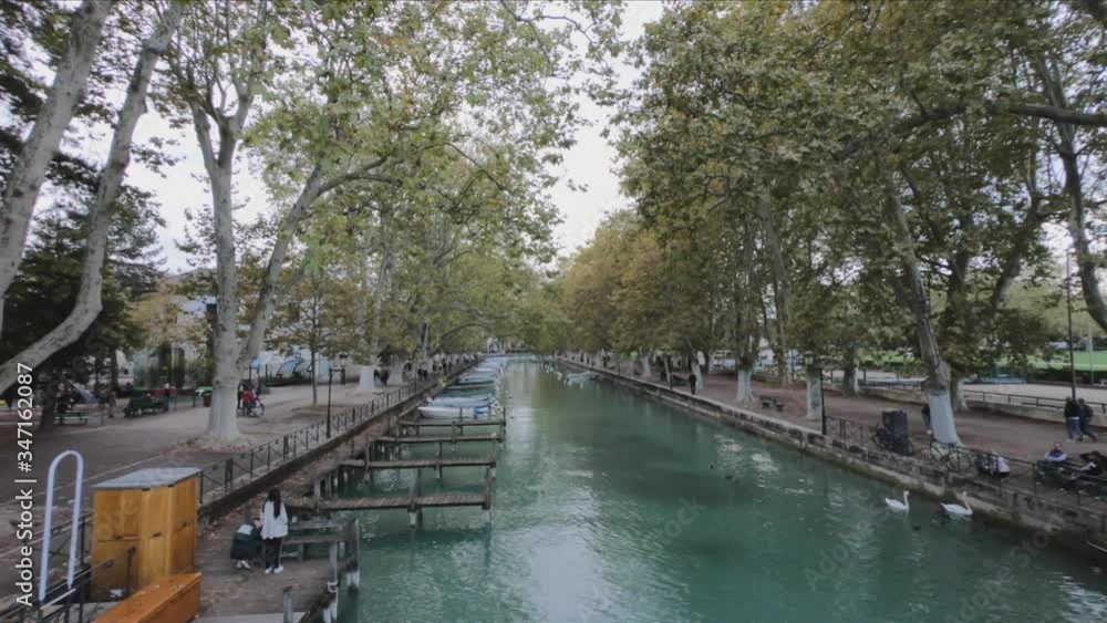PAN SHOT SLOW MOTION - Vassé canal and its moored boats, banks and lines of plane trees in autumn. Canal du Vasse as seen from Pont des Amours, Annecy, France. PAN SHOT SLOW MOTION - Vassé canal and its moored boats, banks and lines of plane trees in autumn. Canal du Vasse as seen from Pont des Amours, Annecy, France.