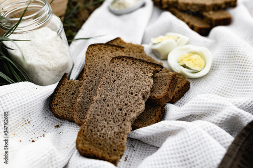 Slices of black bread and a boiled chicken egg on a white towel on the green grass. Picnic in nature with simple food. Next to the salt and flowers with plants in a wooden box.