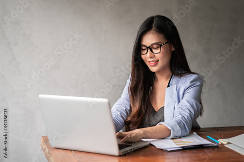 Young happy Asian businesswoman in blue shirt working from home and use a computer laptop and thinking idea for her business