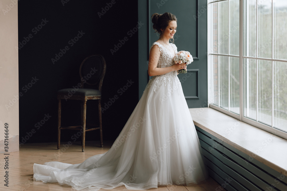 A bride of European appearance, young, is standing face in a wedding dress with a train. Full height photo indoors, front view.