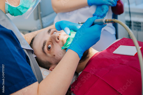 A dentist in blue gloves is drilling the teeth of a young guy in a dental office. A man is afraid of a doctor and wants to scream. Dental Clinic Workflow