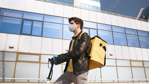Delivery man with yellow bagpack and protective mask standing with electric scooter on business center background, low angle view