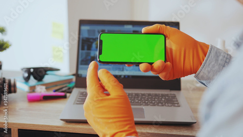 NEW YORK - March, 28, 2020: Young man reading on smartphone greenscreen app holding in horizontal positing sit by desk laptop wokrplace at home. People, technology. Health care. photo