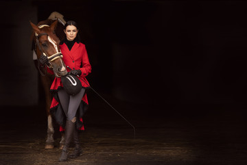 Rider jockey woman in red frock coat holds horse by bridle, fashion photo black background