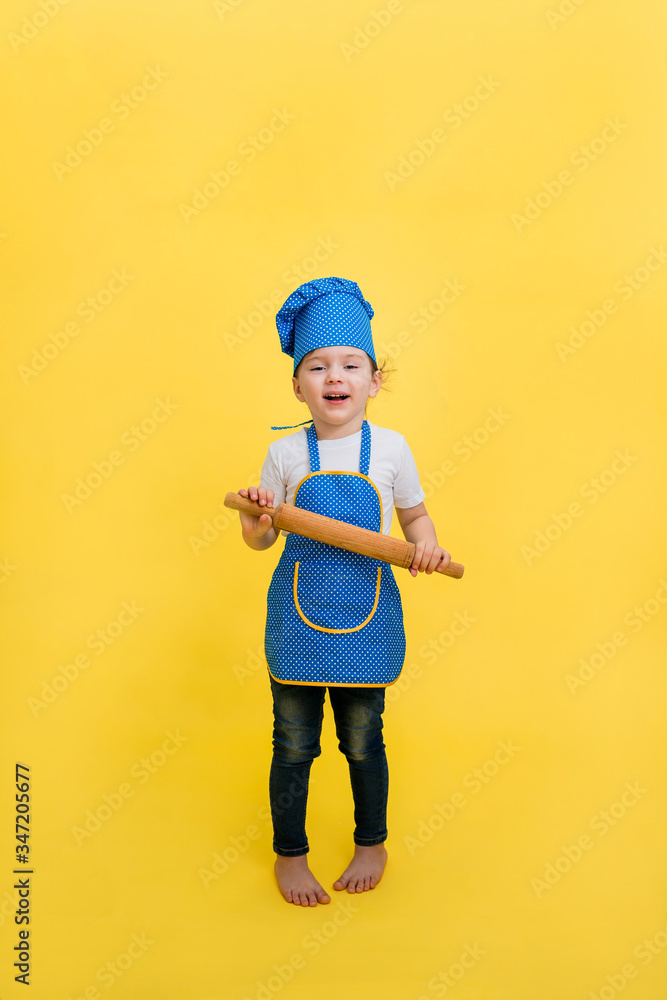 A little girl cook with a rolling pin on a yellow background with an empty seat. A beautiful girl in a full-length apron and hat. A little girl cook with a rolling pin on a yellow background with an empty seat. A beautiful girl in a full-length apron and hat.