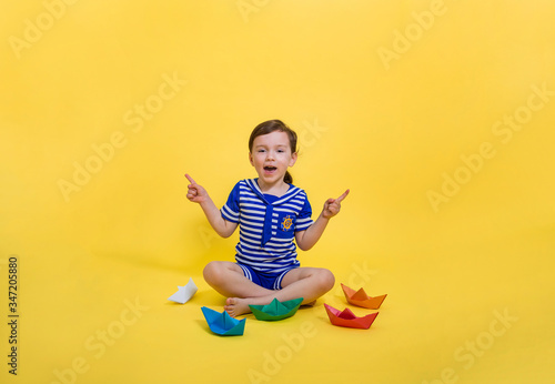 A beautiful sailor girl points her fingers to the side and sits on a yellow isolated background. The girl made origami out of paper. Colorful paper boats.