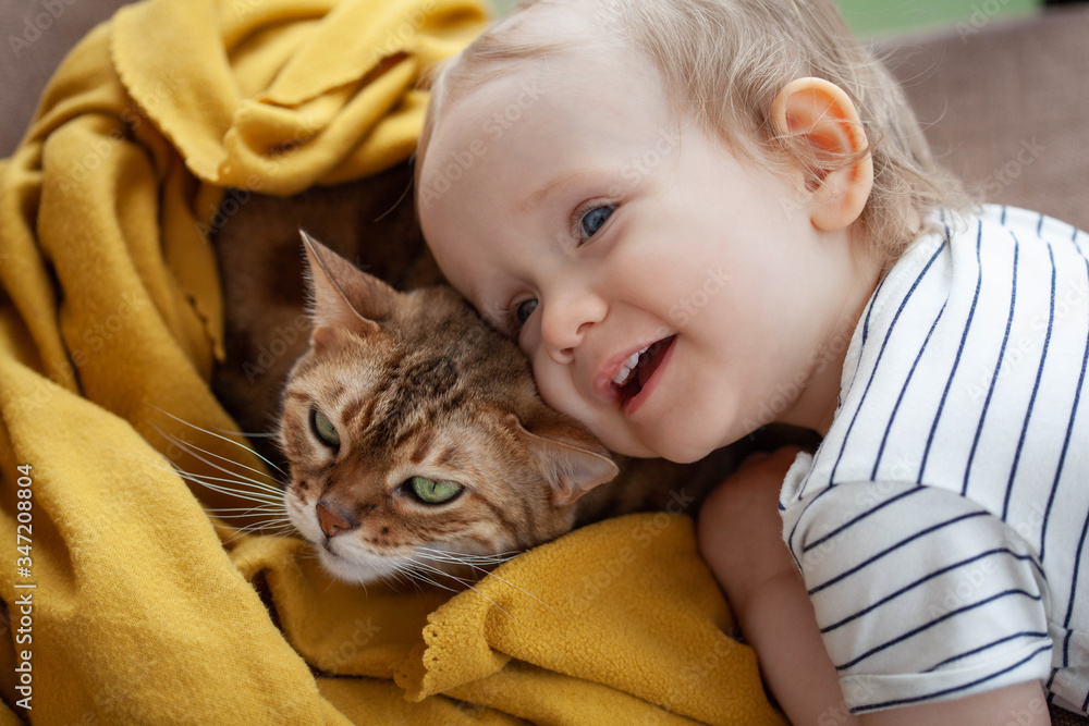 baby girl is hugging a sleeping bengal cat in yellow blanket