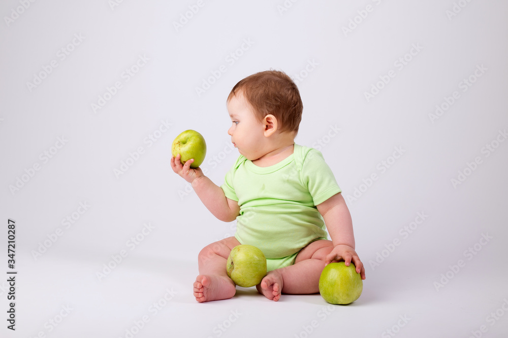 Cute toddler in green bodysuit sits with green apples on white background Cute toddler in green bodysuit sits with green apples on white background