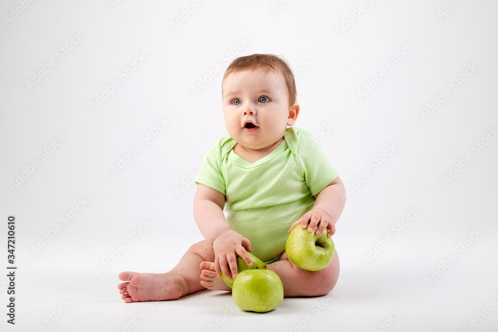 Cute toddler in green bodysuit sits with green apples on white background Cute toddler in green bodysuit sits with green apples on white background