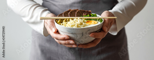 Female hands hold a bowl with Asian traditional soup. Beef pho bo with eggs. Two chopsticks on top.