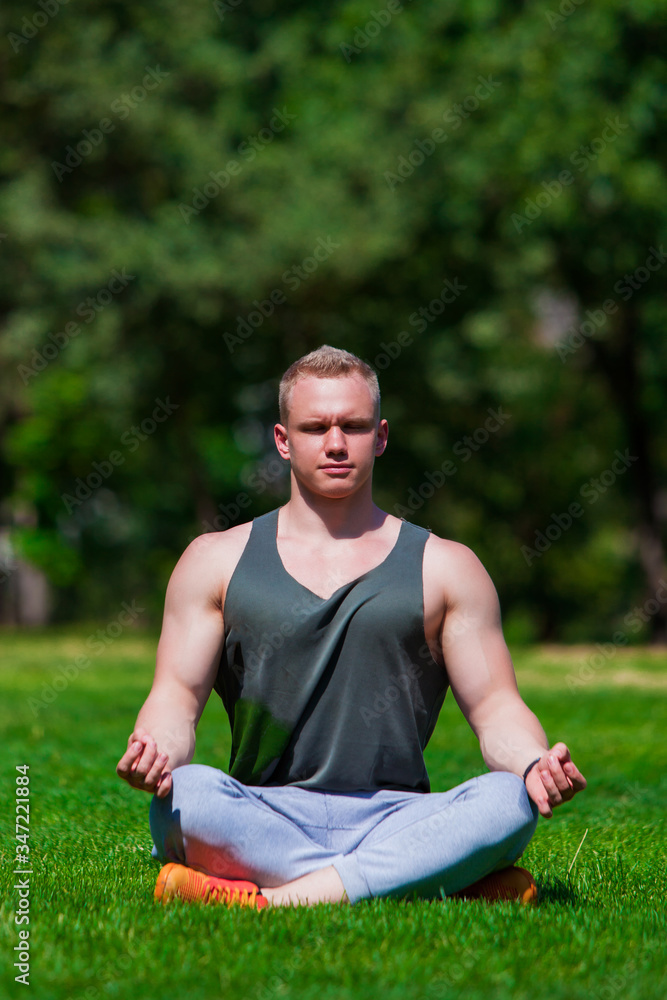 Obraz premium Young muscular man is doing yoga in a summer park. Sporty man is sitting in the Lotus position and relaxing outdoors