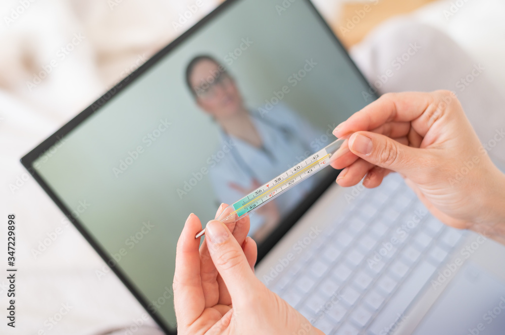 A woman is lying in bed with heat and is holding a thermometer. A friendly doctor talks to a sick patient by video link. Therapist on a laptop screen. Online medical consultation during the quarantine A woman is lying in bed with heat and is holding a thermometer. A friendly doctor talks to a sick patient by video link. Therapist on a laptop screen. Online medical consultation during the quarantine