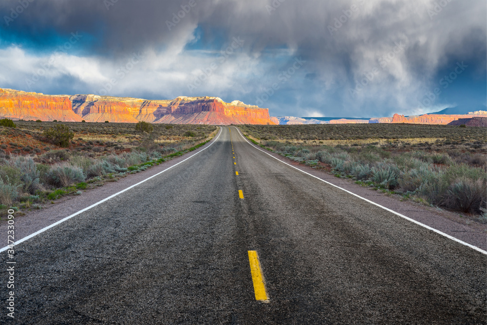 Fototapeta premium The road running through the dry prairie and rests on the red mountains on the background of a stormy sky.