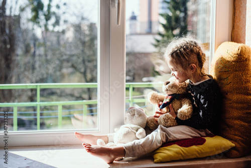 Cute little girl enjoying the sunshine while sitting at the window. The child looks out the window while sitting in the house. Vitamin D. Film noise