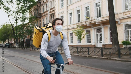 Poartrait of courier with yellow backpack and protective mask rides a bicycle on the street through the city with food delivery and using smartphone photo