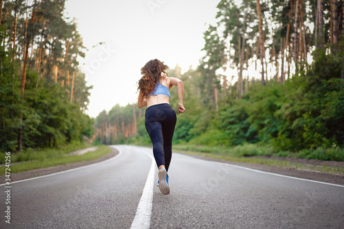 Beautiful caucasian young girl athlete runs sunny summer day on asphalt road in the pine forest view from behind.
