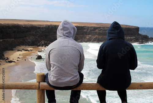Surfer couple sitting on a fence wearing hoodies overlooking the waves hitting the beach and the cliffs