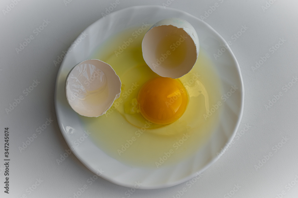 Top down view of one cracked egg with yellow yolk surrounded by egg shells is laying on a white plate on white background Top down view of one cracked egg with yellow yolk surrounded by egg shells is laying on a white plate on white background