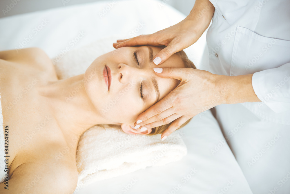 Beautiful happy woman enjoying facial massage with closed eyes in spa salon. Relaxing treatment in medicine and Beauty concept