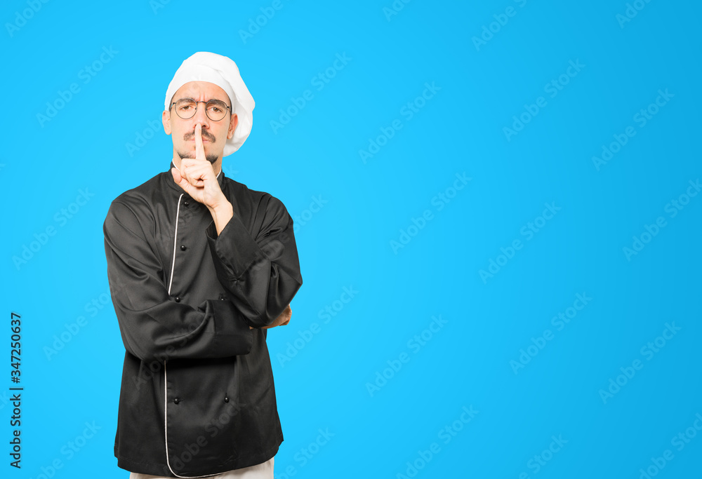 Serious young chef asking for silence gesturing with his finger Serious young chef asking for silence gesturing with his finger