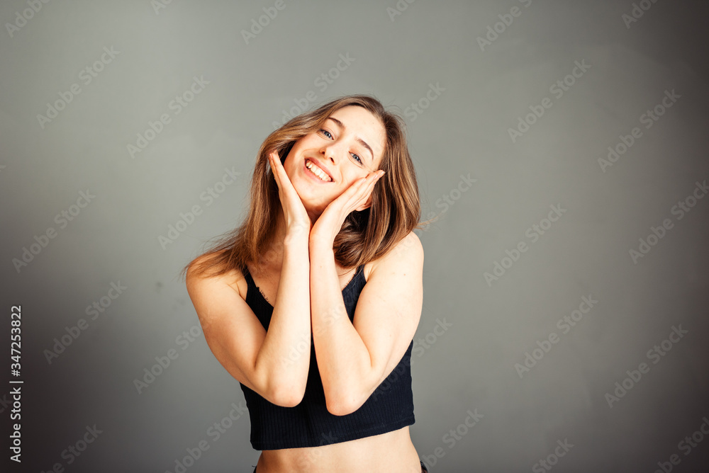 A girl in a black top with her hair loose with hands on her cheeks is smiling on a gray background. Without makeup, without retouching. A girl in a black top with her hair loose with hands on her cheeks is smiling on a gray background. Without makeup, without retouching.