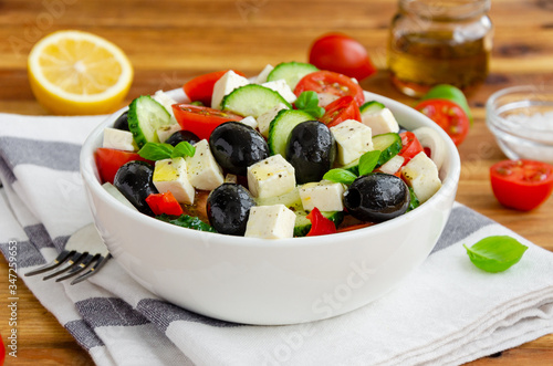 Greek salad of fresh juicy vegetables, feta cheese, herbs and olives in a white bowl on a wooden background. Healthy food. Horizontal orientation, close up.
