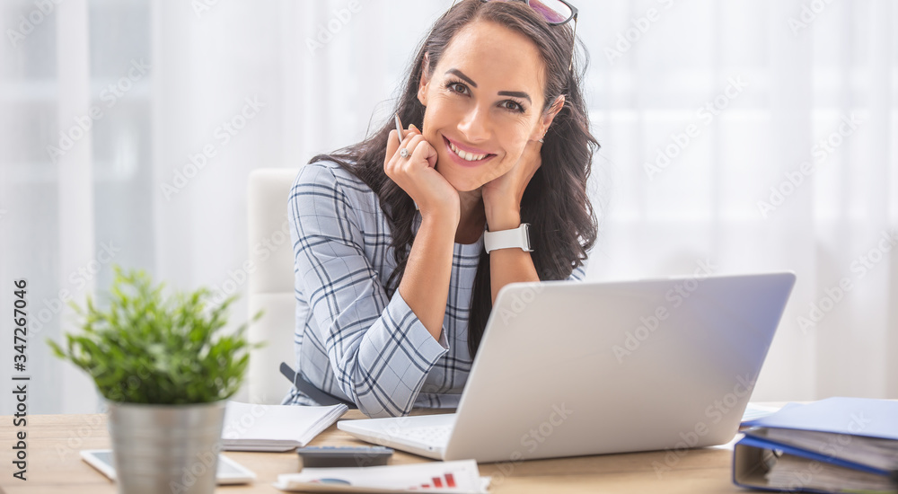 Pretty businesswoman smiles at the camera while sitting at her desk in front of the computer Pretty businesswoman smiles at the camera while sitting at her desk in front of the computer