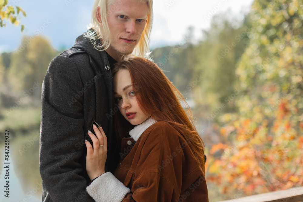 The couple in love in nature cuddle and look into the camera against the backdrop of the river and autumn forest. A young albino guy and a red-haired girl. The couple in love in nature cuddle and look into the camera against the backdrop of the river and autumn forest. A young albino guy and a red-haired girl.