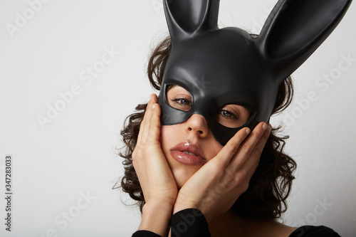 Close-up of young woman with bunny ears mask, raises palm holding her head, gestures over blank space. Isolated.