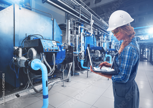 European engineer woman with laptop inspect modern industrial gas boiler room. Heating gas boilers, pipelines, valves. Blue toning with sunflare. Mixed media