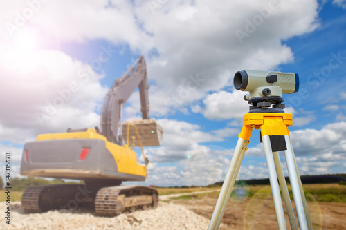 Optical level next to excavator. Concept - an excavator clears places for construction. Preparation of site for construction of the house. Optical level on the sky background. Gyodetic preparation. photo