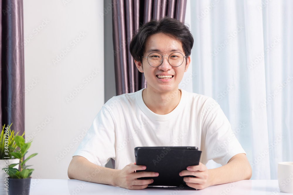 Happy Asian young business handsome man work from home office wear glasses, t-shirt comfortable he smiling and using a black modern smart digital tablet computer to read email or learning on desk Happy Asian young business handsome man work from home office wear glasses, t-shirt comfortable he smiling and using a black modern smart digital tablet computer to read email or learning on desk