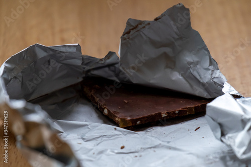 An unwrapped chocolate bar with a corner block of dark chocolate with nuts. The silver wrapper and package is torn open and laying on a wooden table. The sweet dessert snack has antioxidants.  photo