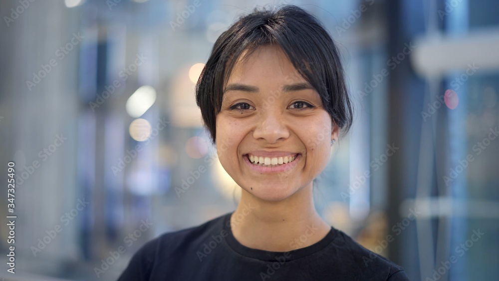 Close up portrait shot of smiling happy black hair asian female with beautiful smile wearing grey sweatshirt standing on background of big mall center. Blue background