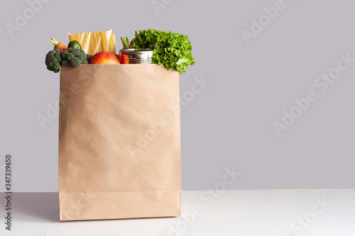 Various grocery items in paper bag on white table opposite gray wall. Bag of food with fresh vegetables, fruits, pasta and canned goods. Food delivery, shopping or donation concept. Copy space.