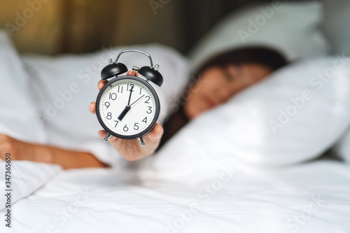 An asian woman holding and showing an alarm clock while sleeping on a white cozy bed in the morning