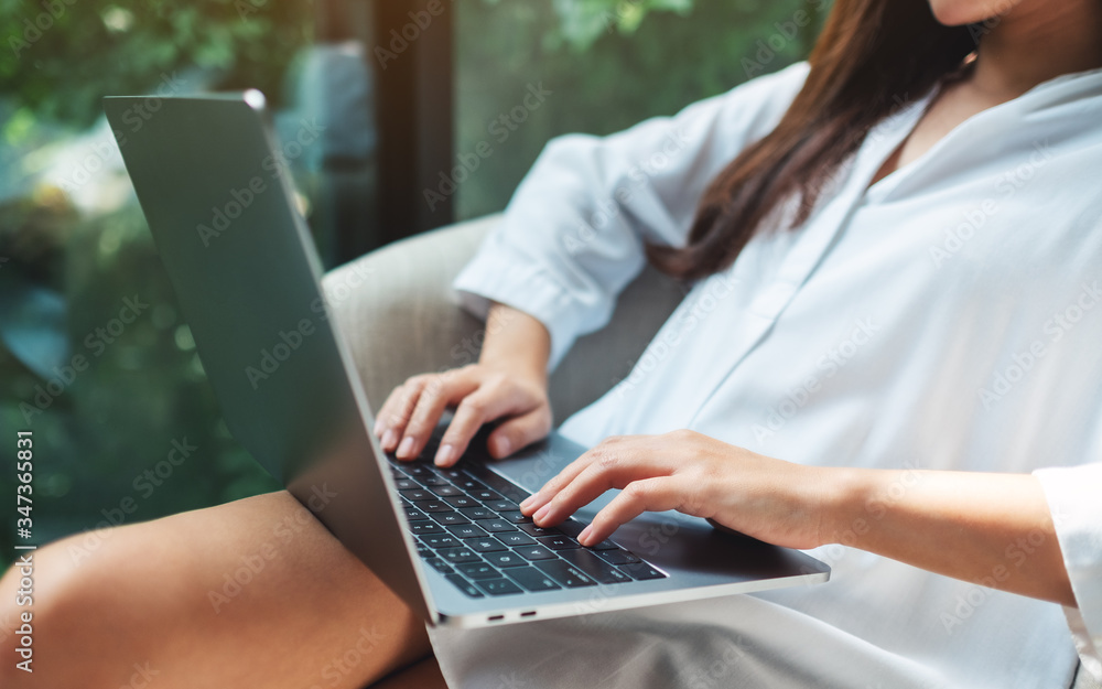 Fototapeta premium Closeup image of a woman working and typing on laptop computer while sitting on a white armchair at home with green nature background