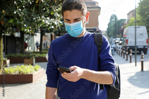 COVID-19 Pandemic Coronavirus Worried Young Man Wearing Surgical Mask Using Smart Phone App in City Street to Aid Contact Tracing and Self Diagnostic in Response to the Coronavirus Pandemic 2019 photo