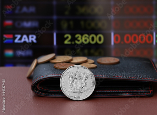The heap coins of South Africa Rand money and black leather wallet on brown floor with digital board of currency exchange money background.