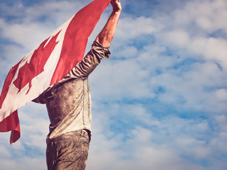 Attractive man holding Canadian Flag on blue sky background on a clear, sunny day. View from the back, close-up. National holiday concept