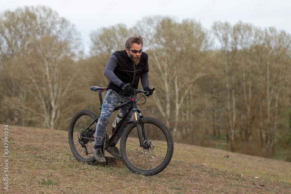 Sports brutal bearded guy on a modern mountain bike. Cyclist on the green hills in the spring.