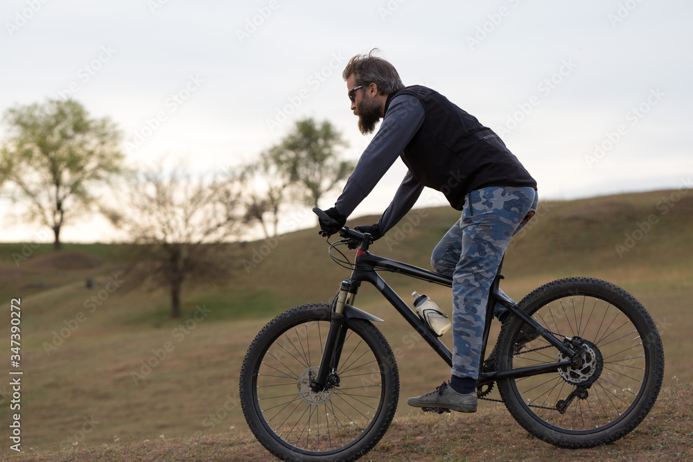 Sports brutal bearded guy on a modern mountain bike. Cyclist on the green hills in the spring.