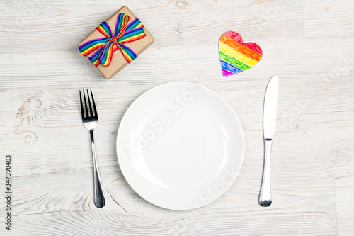 Festive table setting with cutlery and gift with rainbow LGBT ribbon on a light wooden background.