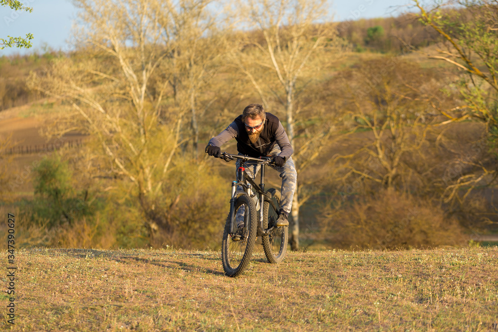 Sports brutal bearded guy on a modern mountain bike. Cyclist on the green hills in the spring.