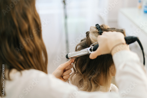 Female hairdresser making hairstyle for young unrecognizable woman with blonde hair in salon, back view, no face. Concept of backstage work.