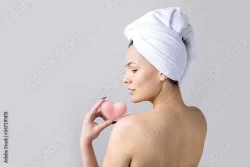 Beauty portrait of woman in white towel on head with a sponge for a body in view of a pink heart. Skincare cleansing eco organic cosmetic spa relax concept.