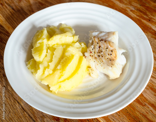 cooked baked cod fish with mashed potatoes on white plate on old wooden table in home kitchen